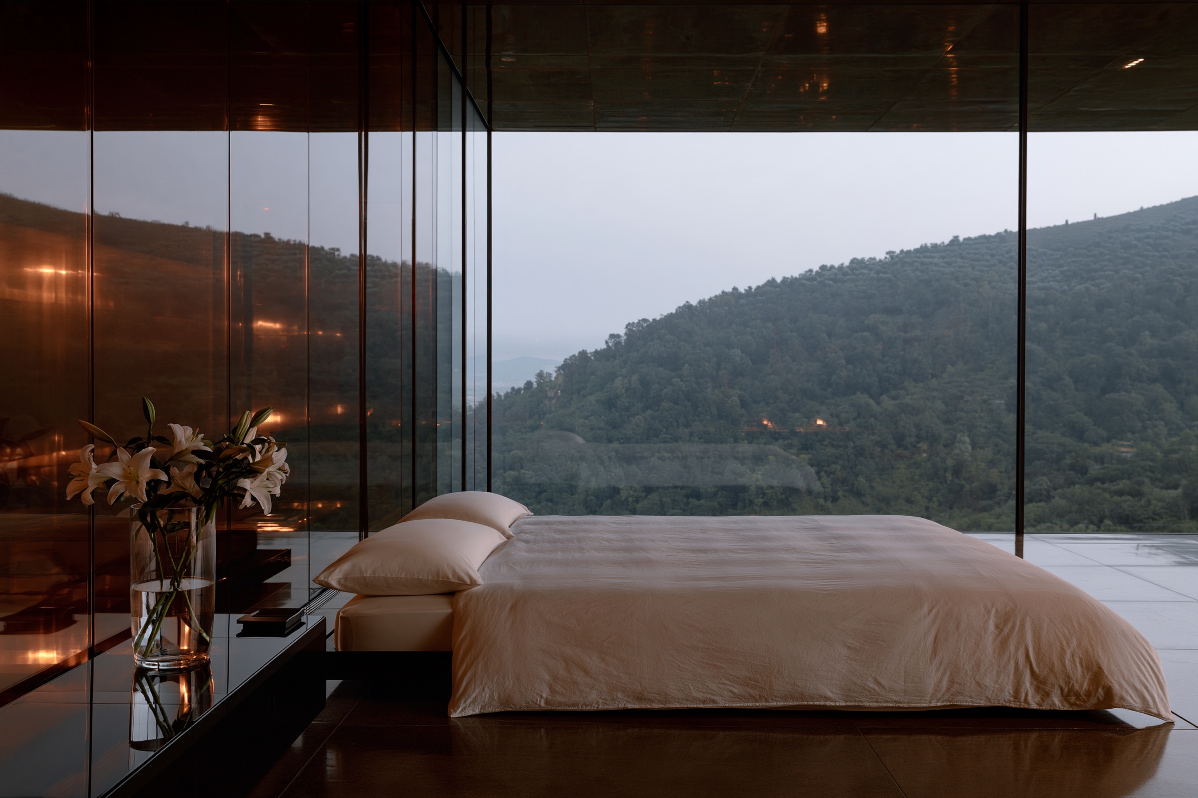 A guest room at dusk, a low pale bed and lamplight, the hills beyond the glass