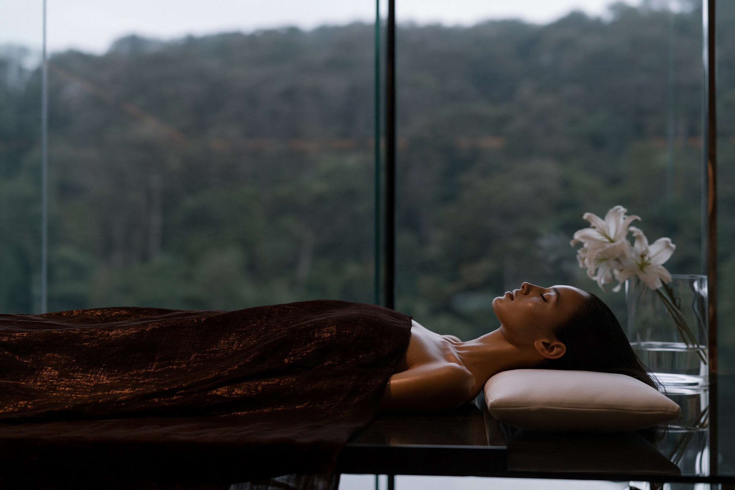 A woman resting under a heavy throw on a treatment bed, a glass wall and trees behind her