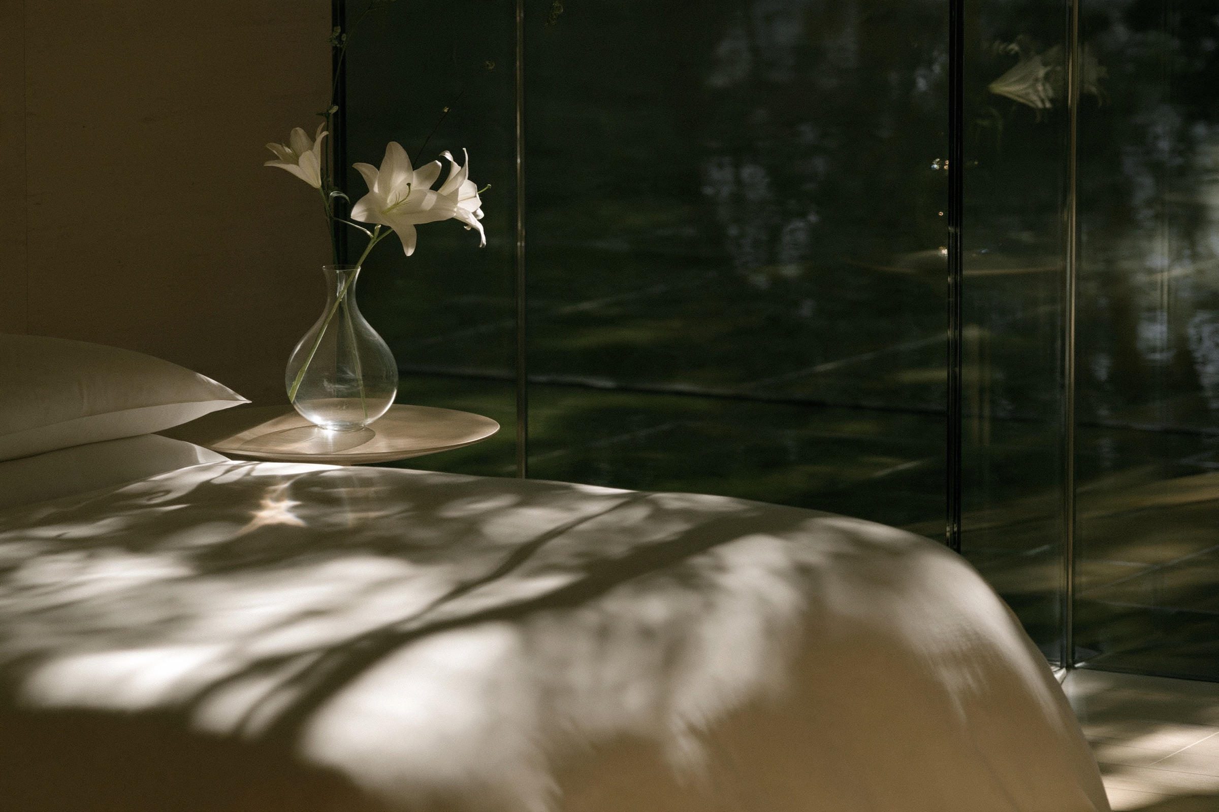 A guest room in daylight, a white bed and a lily in a glass vase against green glass
